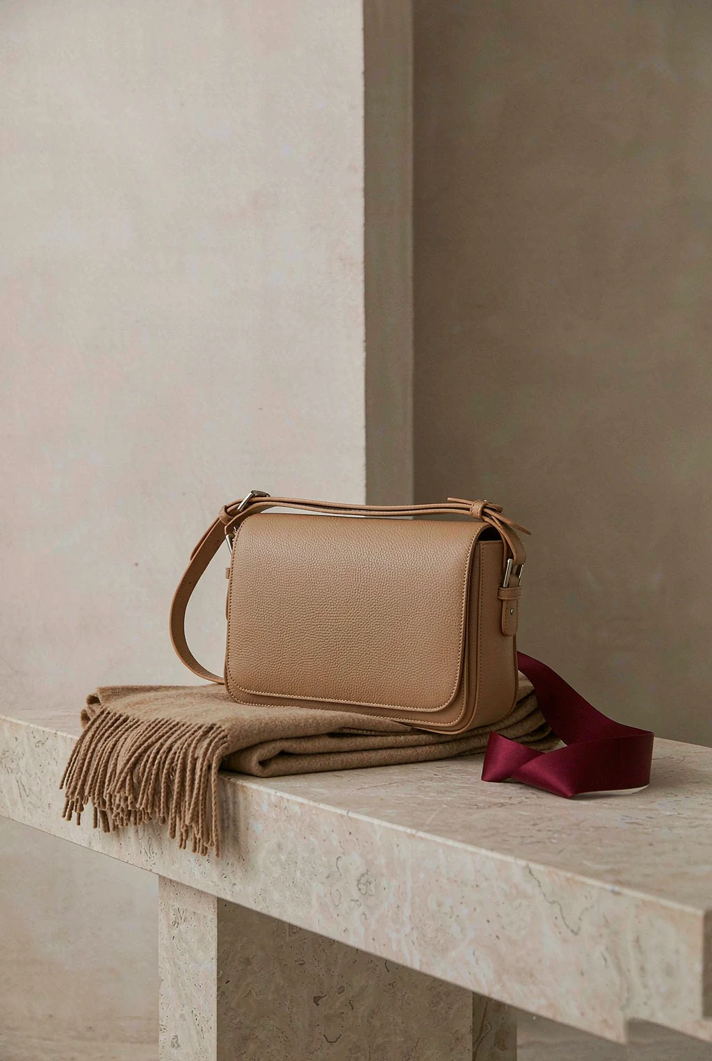 Tan leather satchel on a pale stone bench with a camel wool throw